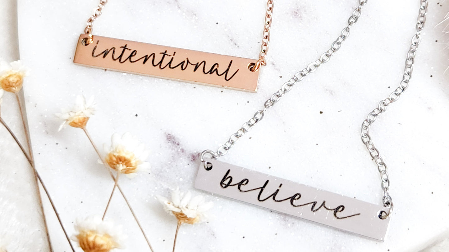 Two necklaces with motivational pendants on a white surface with dried flowers.