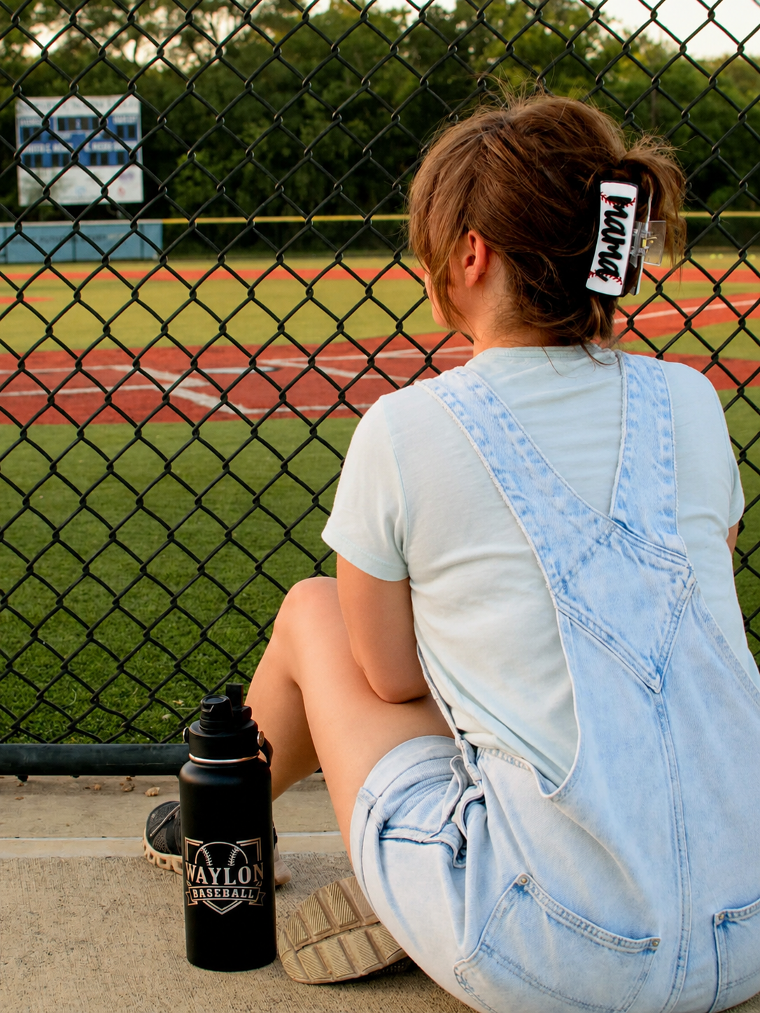 A person sitting on a concrete ledge at a baseball field, seen from behind, wearing a light blue denim overall with custom claw clip baseball mama and a white t-shirt. A black water bottle with 'MAYLON' engraved on it sits next to them.